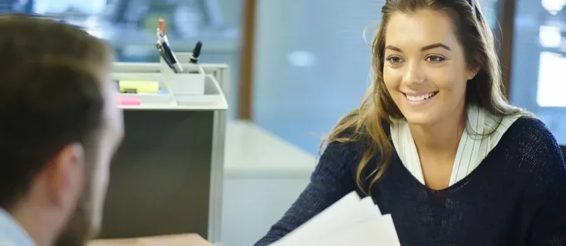 A young female office worker chats to her supervisor across a desk in a busy office.